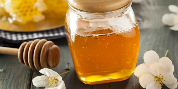 A clear glass jar filled with golden honey and topped with a wooden lid, placed on a dark rustic table alongside a wooden honey dipper and white blossoms.