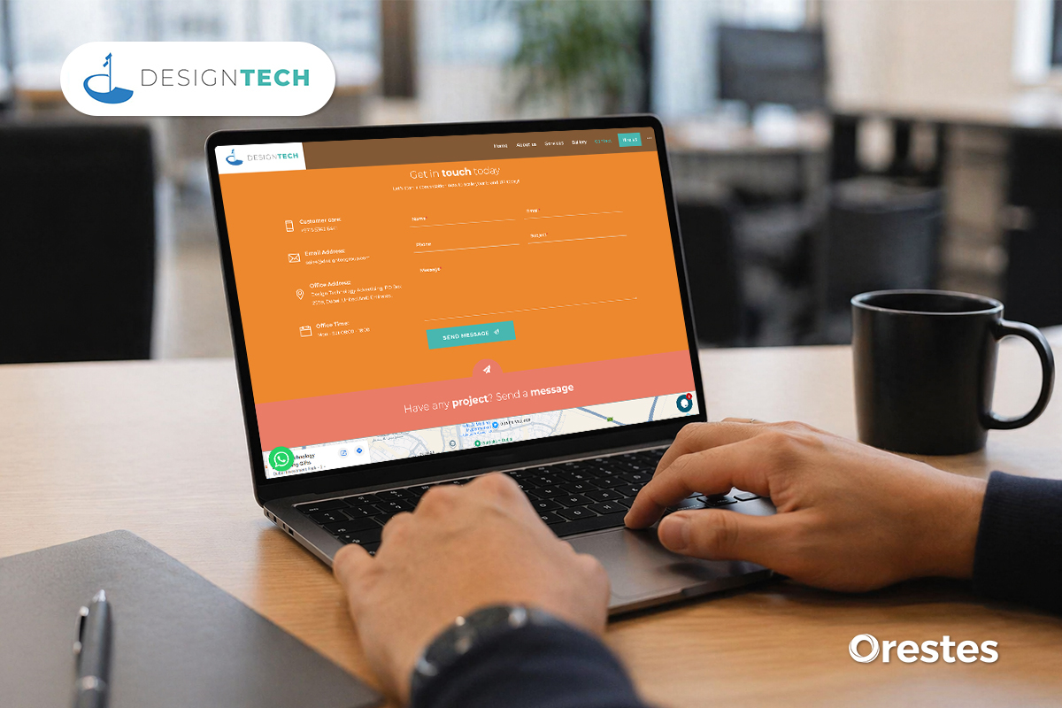 A large desktop monitor on a wooden desk in a bright room with a view of greenery outside. The screen displays the Design Tech "Design & Creative Agency" homepage with cards for Designing, Signage, and Installation.