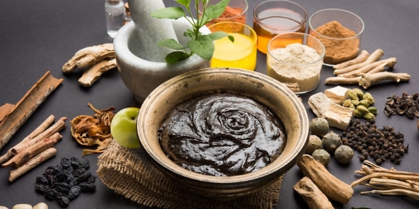 High-angle close-up of a central bowl of Ayurvedic paste, amla, and a white mortar with herbs, surrounded by medicinal roots and spices on a dark surface.