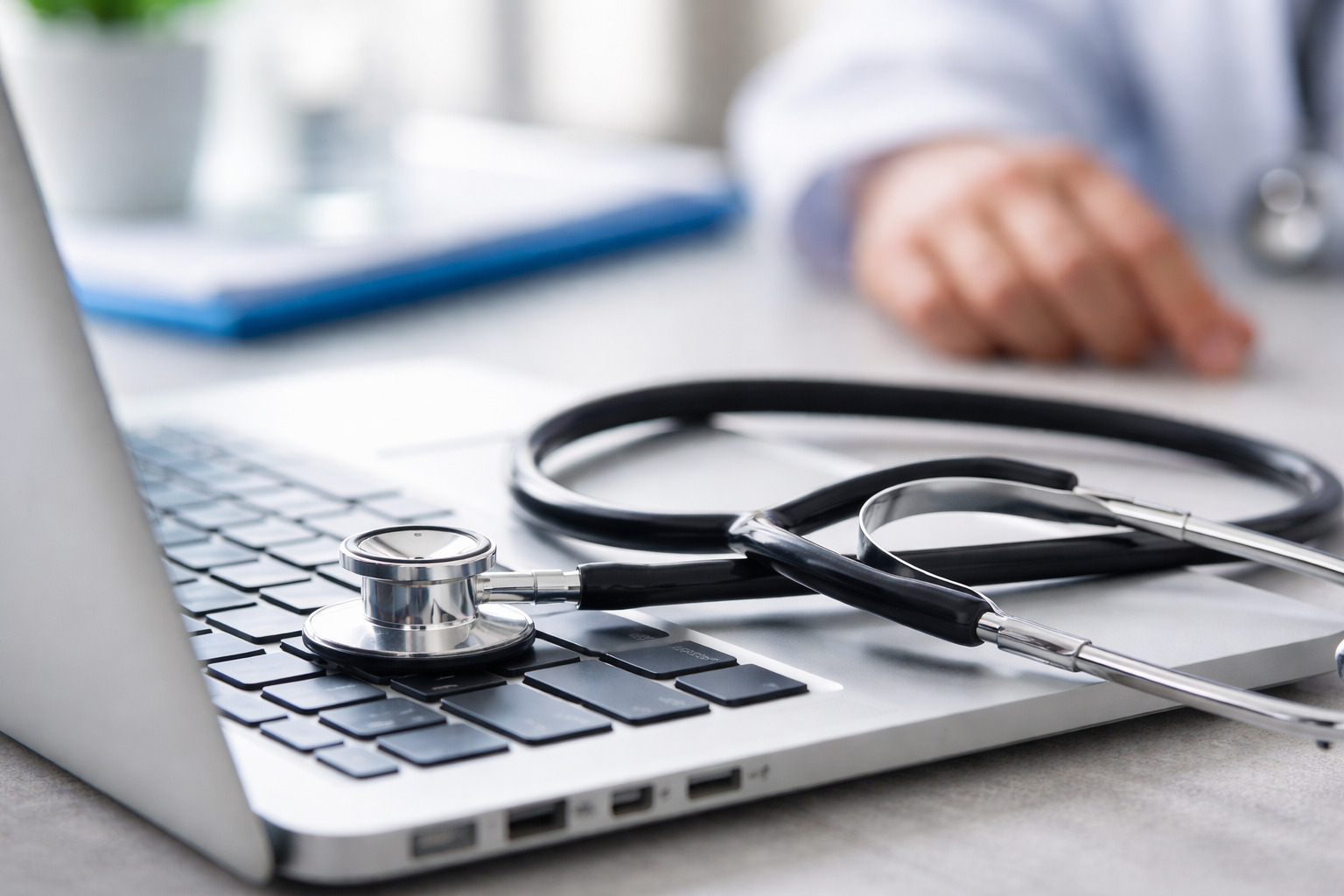 A stethoscope resting on a laptop keyboard with a doctor working in the background, symbolizing the intersection of modern technology and healthcare in today's digital medical practice.
