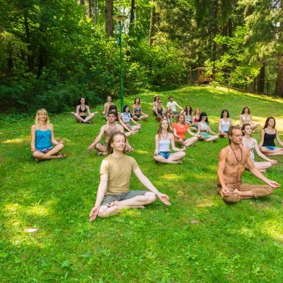 A group of people sitting cross-legged in a meditative yoga pose on a lush green grass lawn surrounded by trees in a forest or park setting.